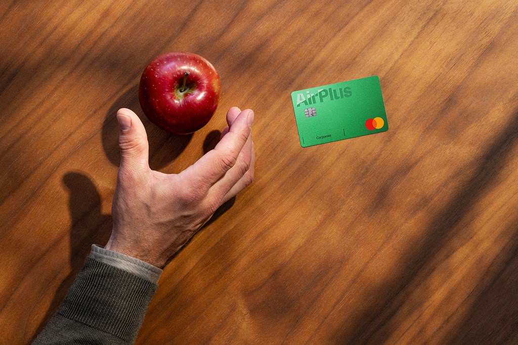En hånd hviler på en trebordflate nær et rødt eple, med et grønt AirPlus Corporate Card ved siden av.