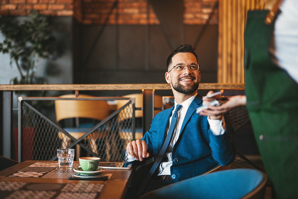 A smiling man in a blue suit sits at a café table and makes a contactless payment to a waiter.