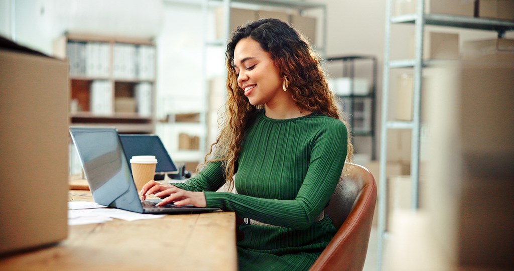 Smiling woman in a green sweater works on a laptop at a desk with a coffee cup in an office.