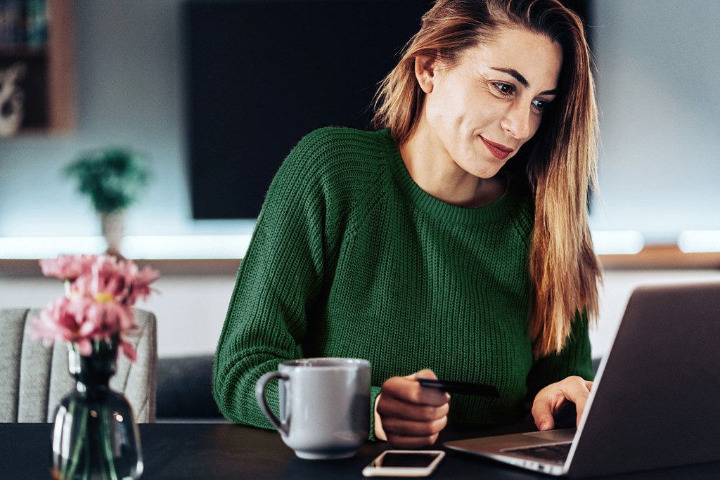 Smiling woman in a green sweater sits at a desk with a laptop and coffee cup, making an online payment.