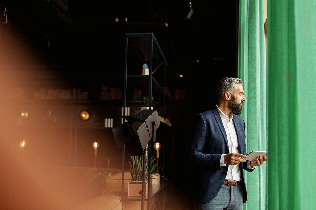 Man in a dark blue blazer holding a tablet stands by a window with green curtains, looking outside.