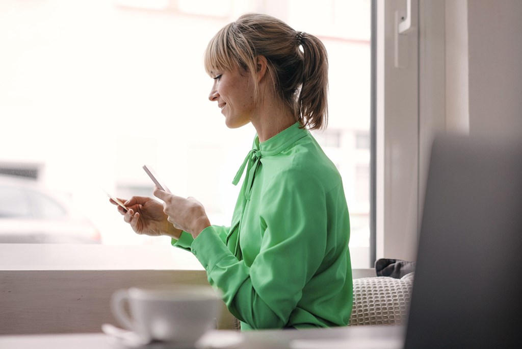 Smiling woman in a green blouse sits by a window holding a mobile phone and a credit card.