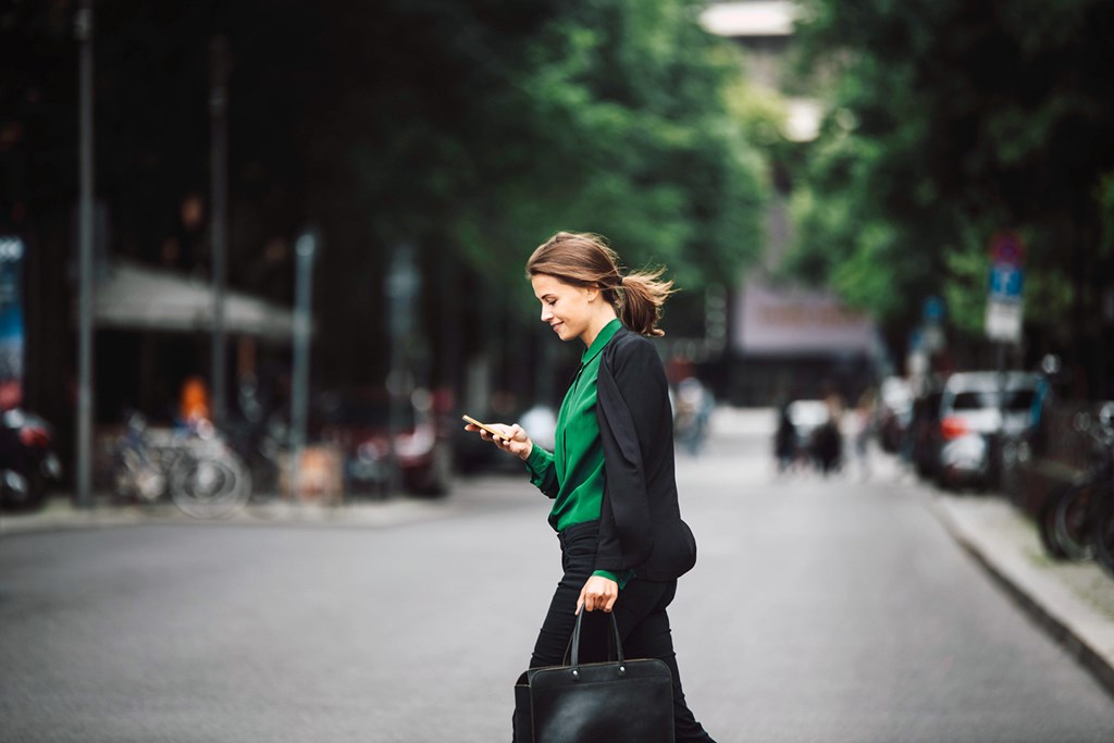 Woman in a green blouse walking outdoors looking at her phone while holding a black handbag.