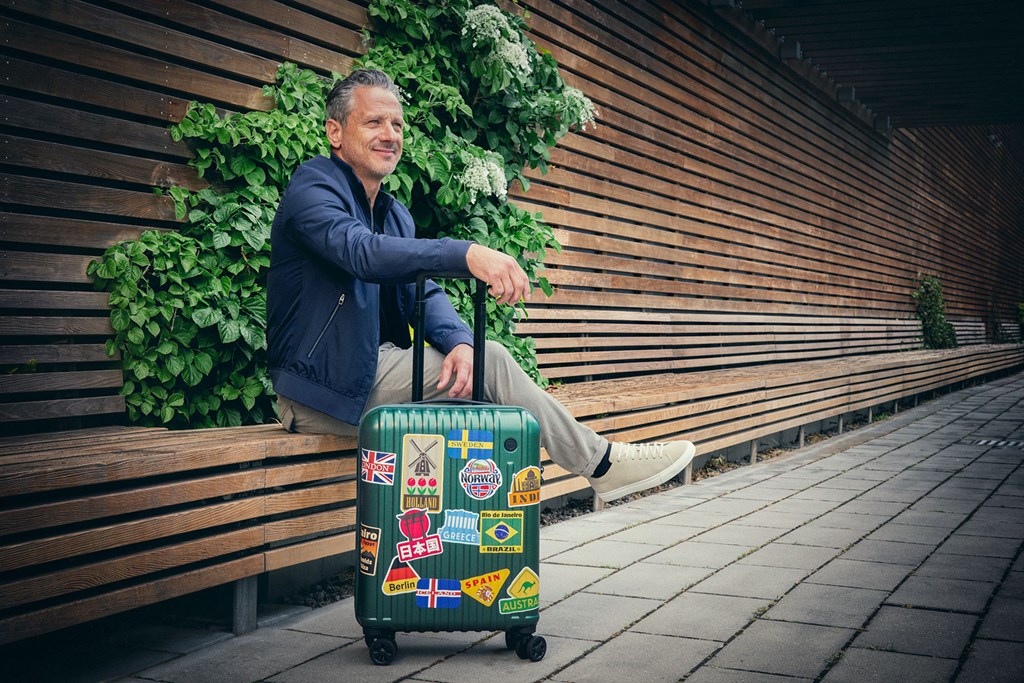 Smilende mann sitter på en benk med en koffert dekket av reiseklistremerker ved en trevegg.