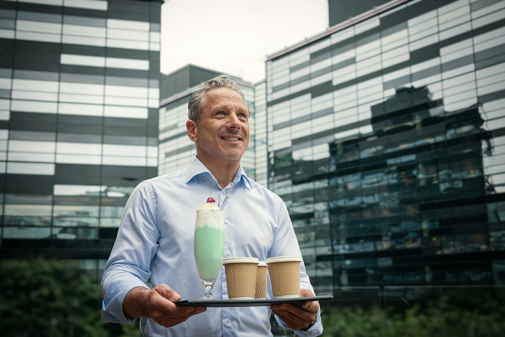 Lächelnder Mann im hellblauen Hemd hält ein Tablett mit zwei Kaffeebechern und einem grünen Milchshake.