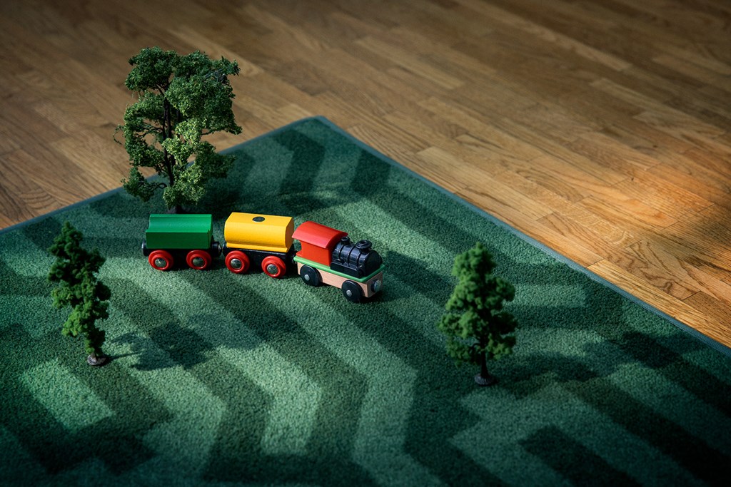 Toy train with red, yellow, and green carriages on a patterned green carpet with small model trees.
